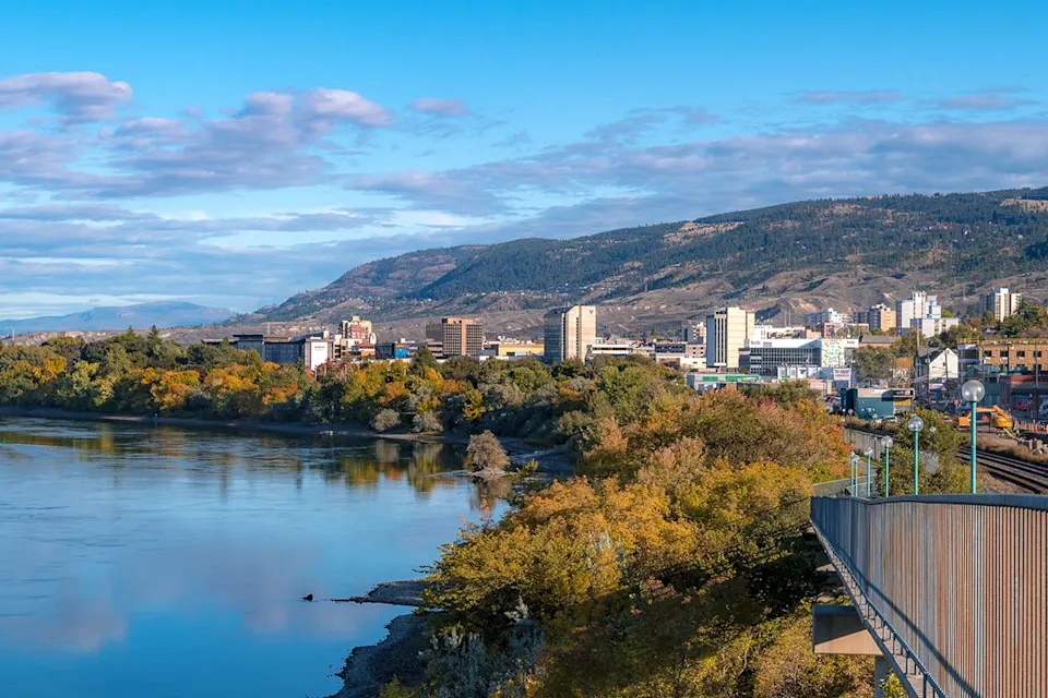 View of Kamloops cityscape with a river, greenery, and mountains in the background on a partly cloudy day.
