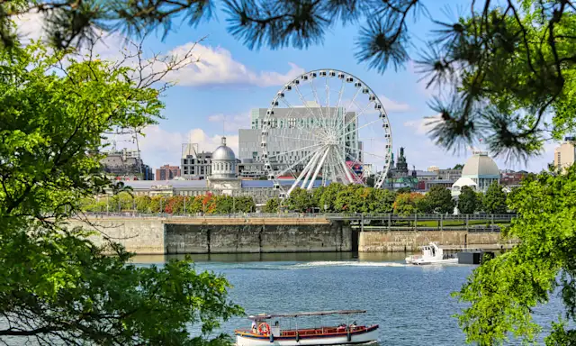 Montreal waterfront with Ferris wheel, historic buildings, and boat on a sunny day framed by green trees.