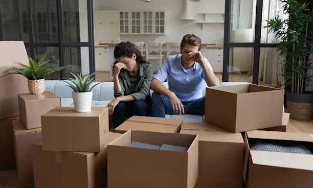 Stressed couple sitting on a couch surrounded by moving boxes in a new home.