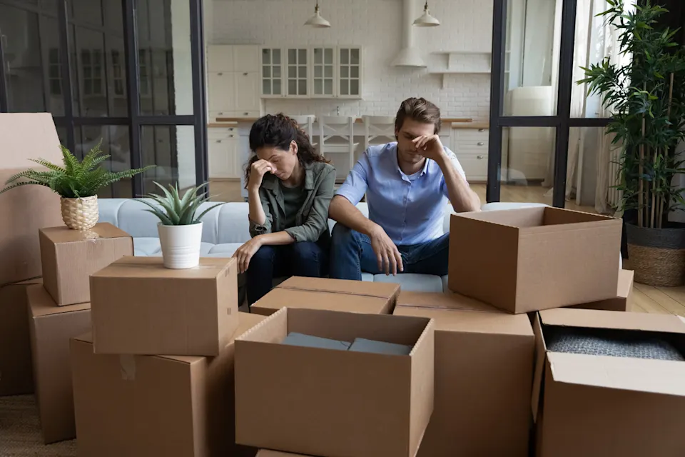 Stressed couple sitting on a couch surrounded by moving boxes in a new home.