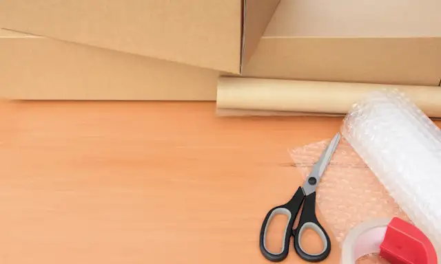 Packing supplies on a wooden surface, including cardboard boxes, scissors, bubble wrap, tape, and packing paper.