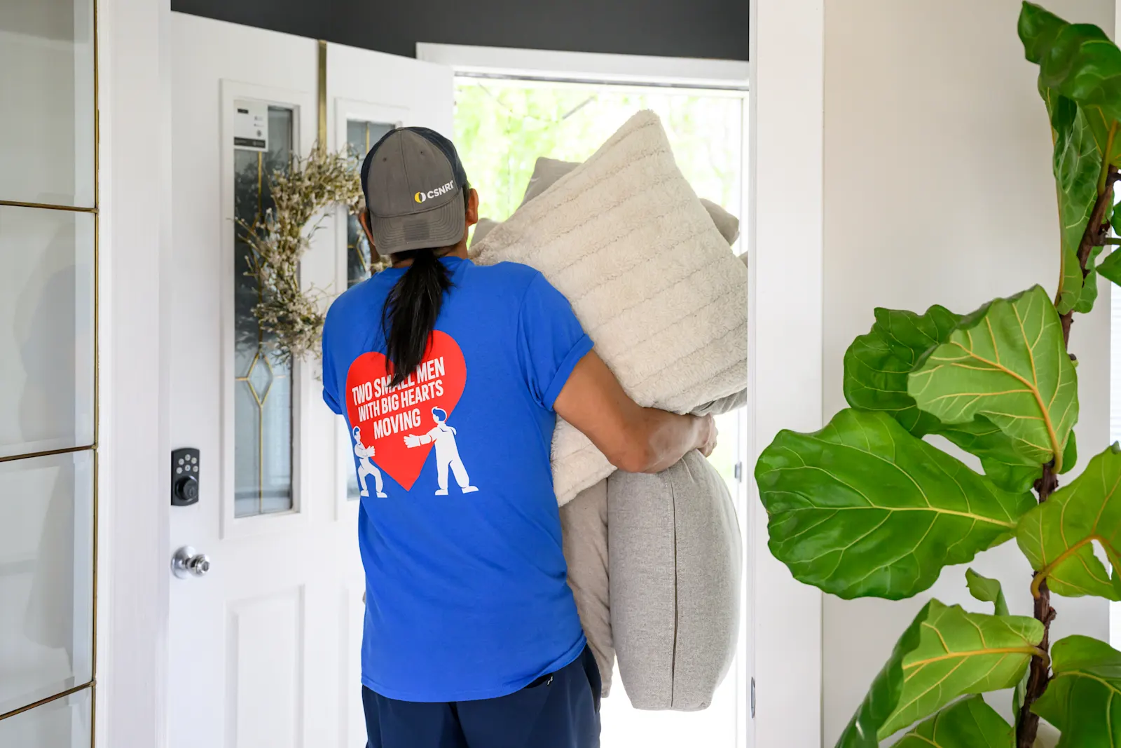 Man in a Two Small Men with Big Hearts Moving shirt carrying pillows into a house.