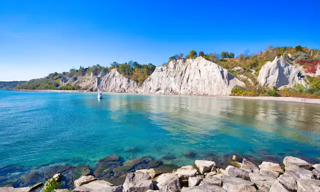 Scarborough Bluffs in Toronto, Ontario, with clear blue water, rocky shoreline, and a sailboat on a sunny day.