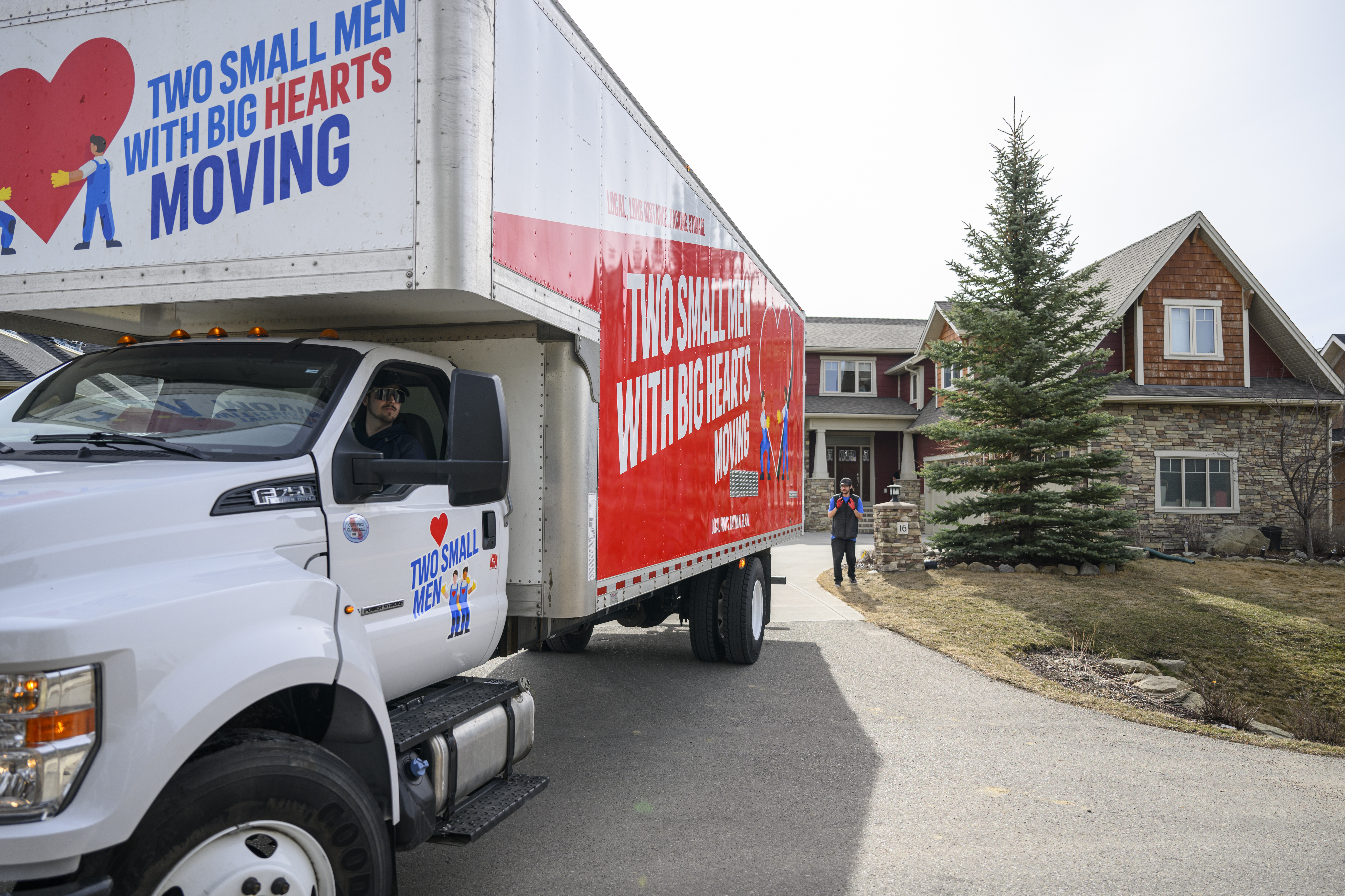 Two Small Men with Big Hearts Moving truck being backed up towards a house, with one mover guiding the truck and another in the driver's seat.