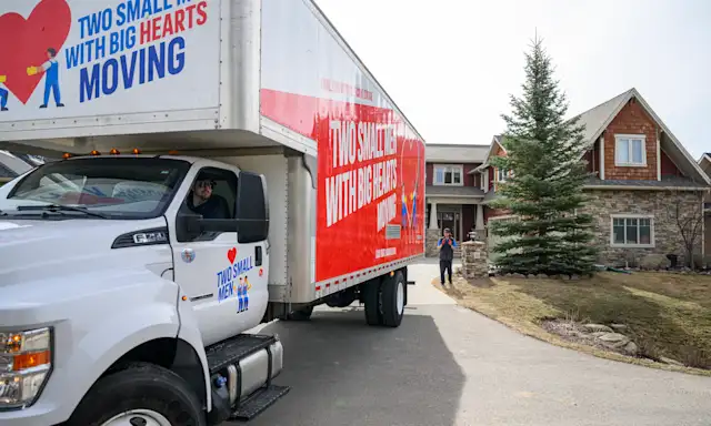 Two Small Men with Big Hearts Moving truck being backed up towards a house, with one mover guiding the truck and another in the driver's seat.