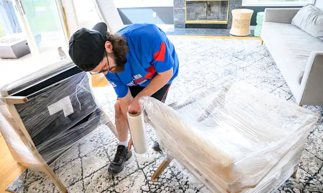 Man in a Two Small Men with Big Hearts Moving shirt wrapping furniture with protective plastic in a living room