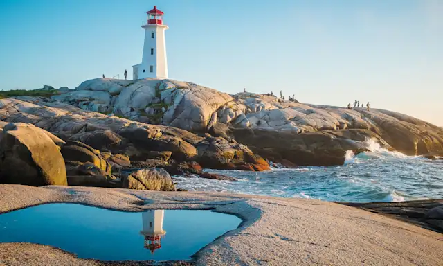 Peggy's Cove Lighthouse in Nova Scotia reflected in a tidal pool on a rocky shoreline during sunset.