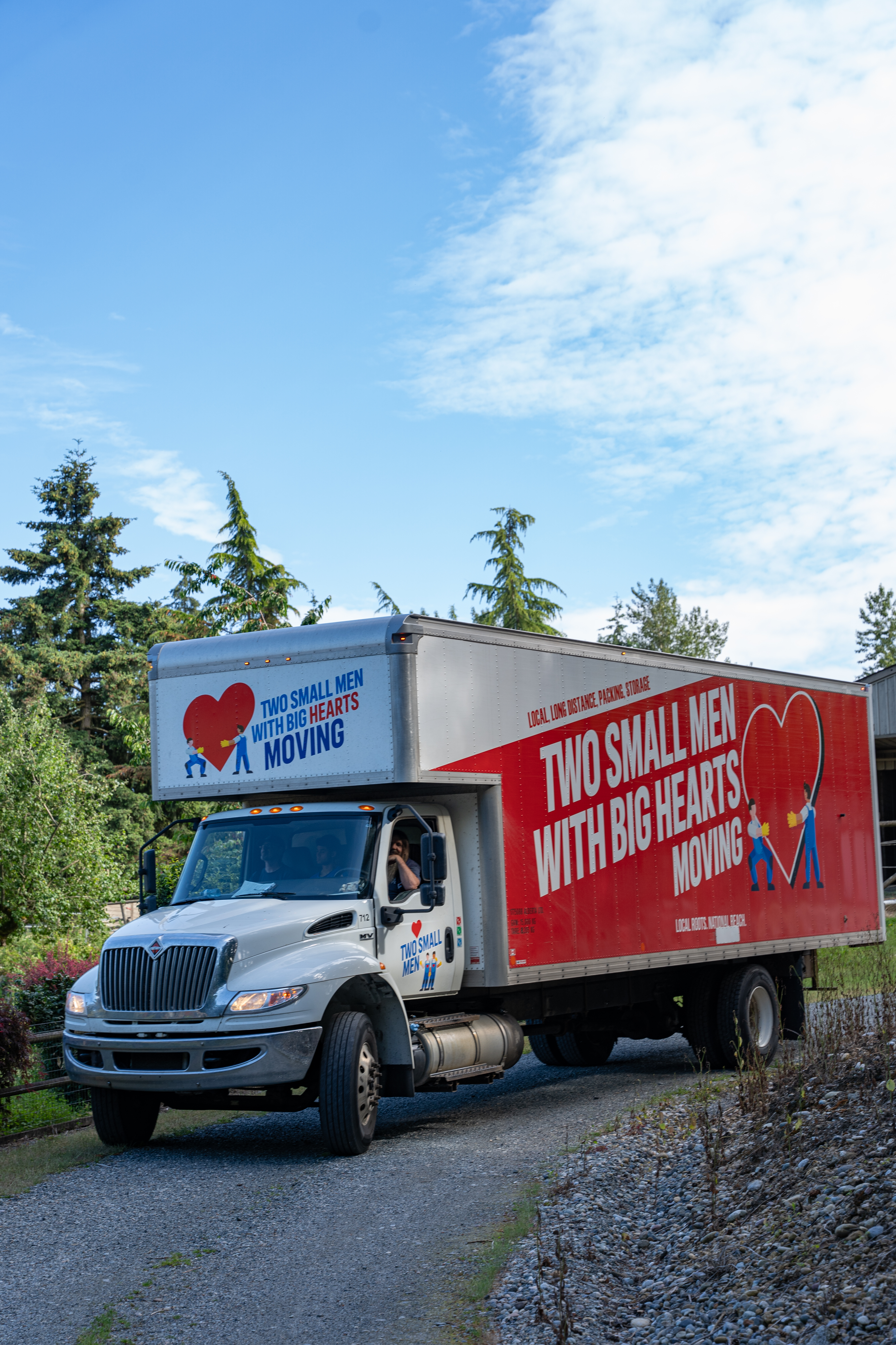A "Two Small Men with Big Hearts Moving" truck is driving down a gravel road, with trees and a clear blue sky in the background.