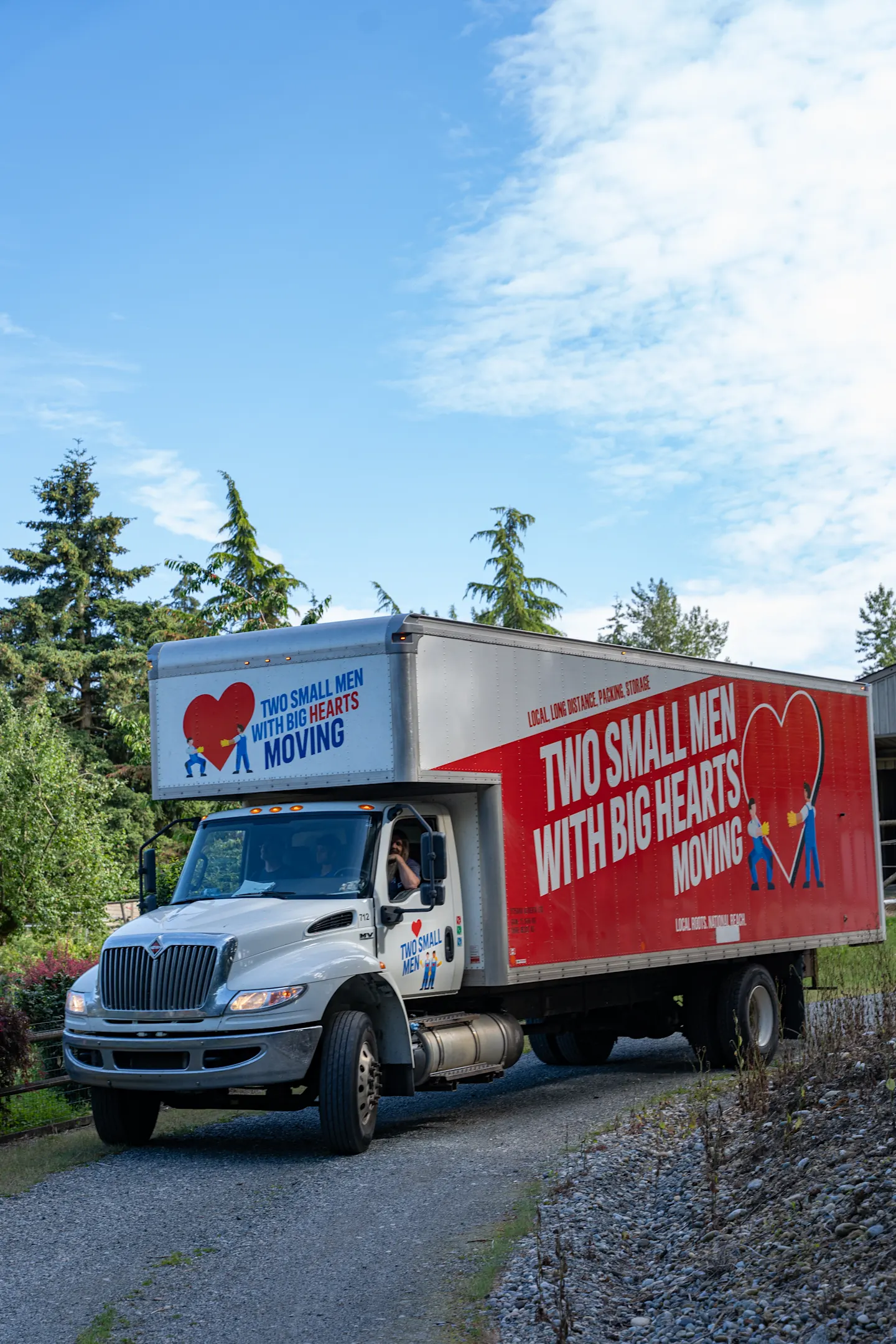 A "Two Small Men with Big Hearts Moving" truck is driving down a gravel road, with trees and a clear blue sky in the background.