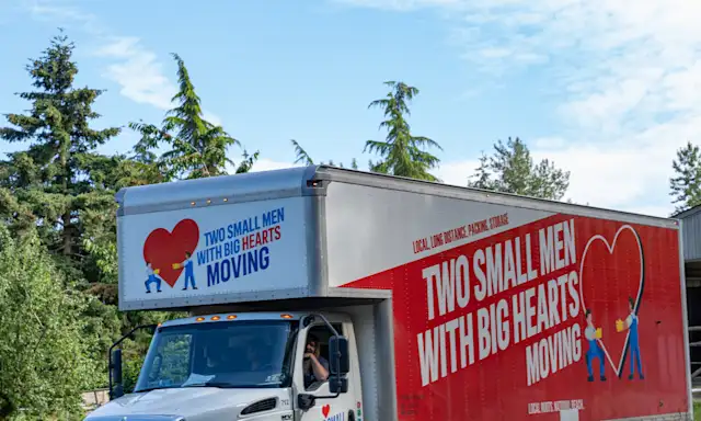 A "Two Small Men with Big Hearts Moving" truck is driving down a gravel road, with trees and a clear blue sky in the background.