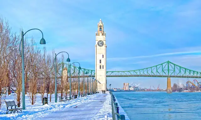 Clock tower along a snowy riverside path with the Jacques Cartier Bridge in the background.