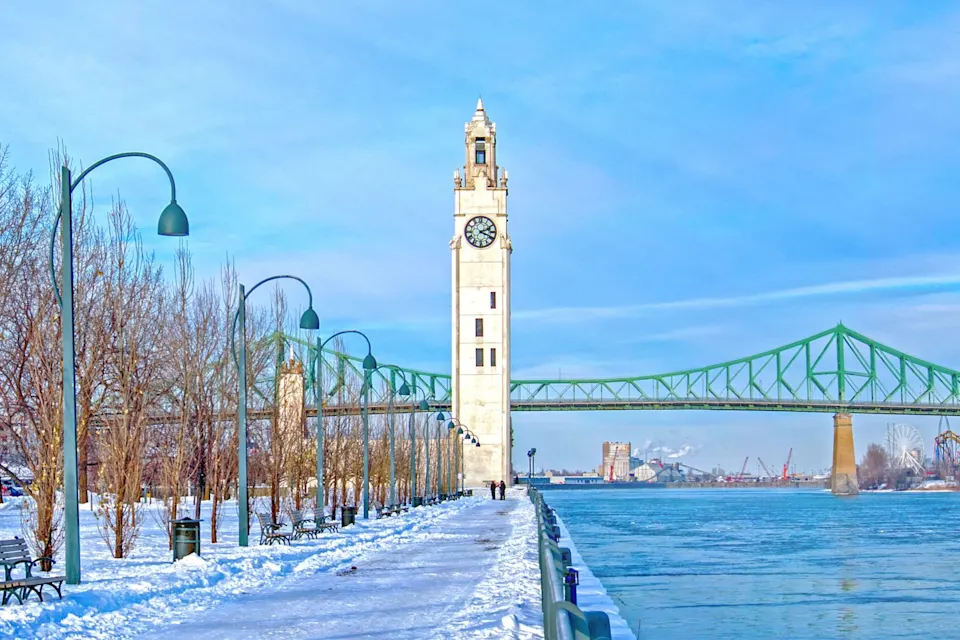 Clock tower along a snowy riverside path with the Jacques Cartier Bridge in the background.