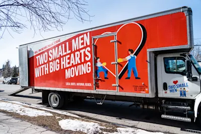 A "Two Small Men with Big Hearts Moving" truck is parked on a street during winter, featuring the company’s logo with cartoon movers and a large heart on its red side. Snow is visible on the ground.