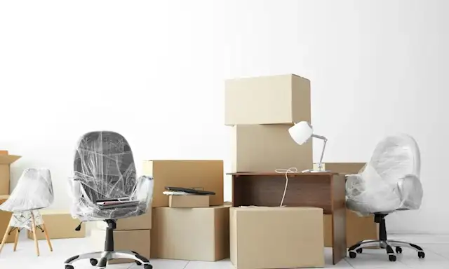 An office space in transition, filled with packed moving boxes, wrapped chairs, and furniture ready for relocation.