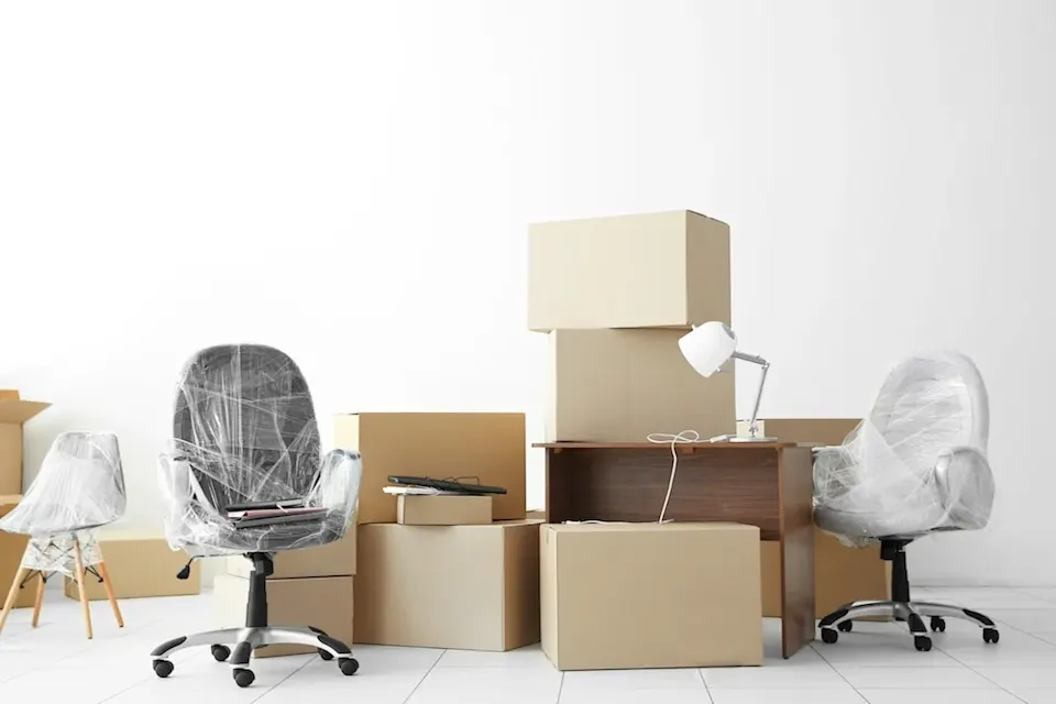 An office space in transition, filled with packed moving boxes, wrapped chairs, and furniture ready for relocation.
