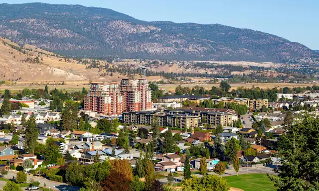 Aerial view of Penticton with residential buildings, greenery, and mountains in the background on a sunny day.