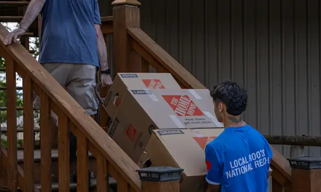 Two movers are carrying large boxes up a wooden staircase, one wearing a "Two Small Men with Big Hearts" shirt. A house with a metal roof is visible in the background.