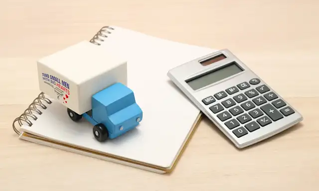A miniature "Two Small Men with Big Hearts" moving truck sits on a notebook next to a calculator, symbolizing moving cost calculations.