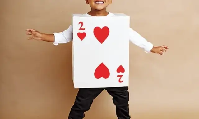 A smiling child is dressed in a "2 of Hearts" playing card costume, posing with arms outstretched against a beige background.
