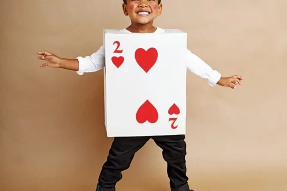 A smiling child is dressed in a "2 of Hearts" playing card costume, posing with arms outstretched against a beige background.