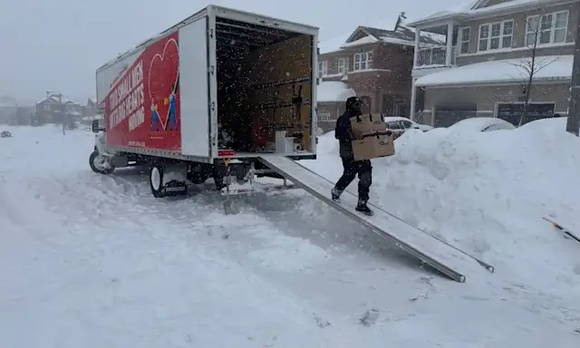 Two Small Men With Big Hearts mover carrying boxes down a truck ramp in heavy snow on a residential street in Toronto