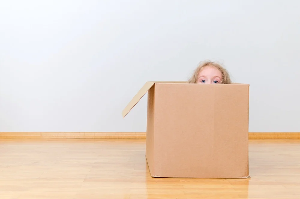 A playful moment during a move, with a child peeking out from inside a large cardboard box. The image captures the joy and lightheartedness of moving day.