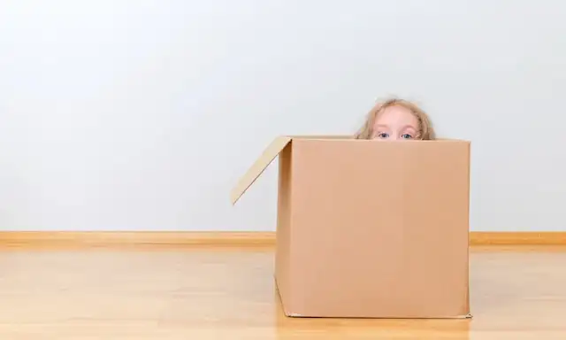 A playful moment during a move, with a child peeking out from inside a large cardboard box. The image captures the joy and lightheartedness of moving day.
