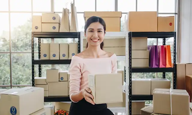 Smiling woman holding a package in a workspace filled with boxes.