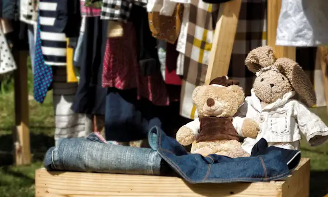 Stuffed bear and bunny toys on display with folded jeans and hanging clothes in the background at an outdoor market or yard sale.