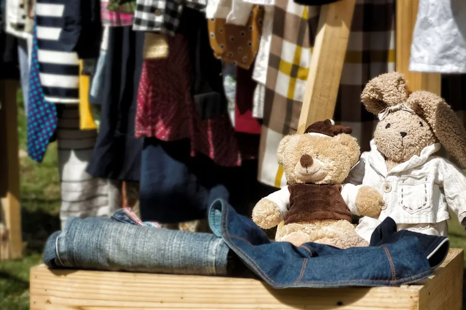 Stuffed bear and bunny toys on display with folded jeans and hanging clothes in the background at an outdoor market or yard sale.