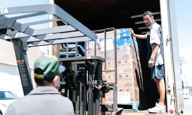 Worker using a forklift to load pallets into a truck while another worker guides the process.