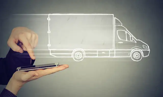 A hand is holding a tablet while another hand interacts with it, displaying a digital outline of a moving truck above the screen, representing digital logistics or fleet management.