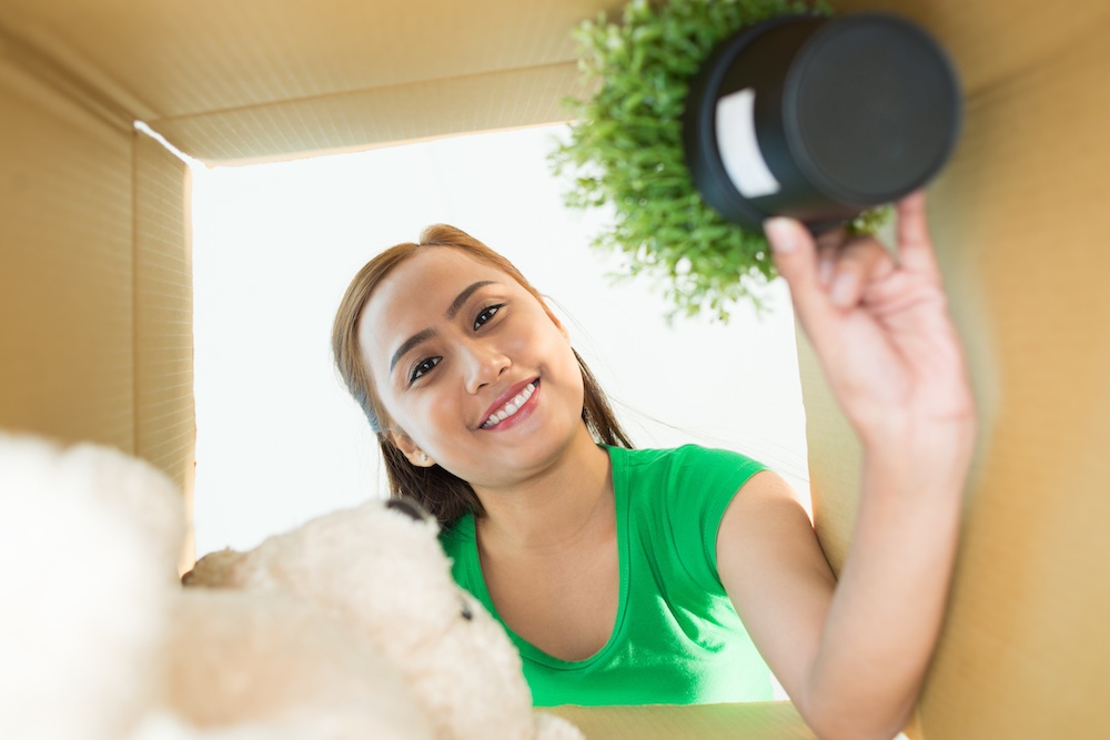 Smiling woman unpacking items from a moving box, including a small plant and a teddy bear.