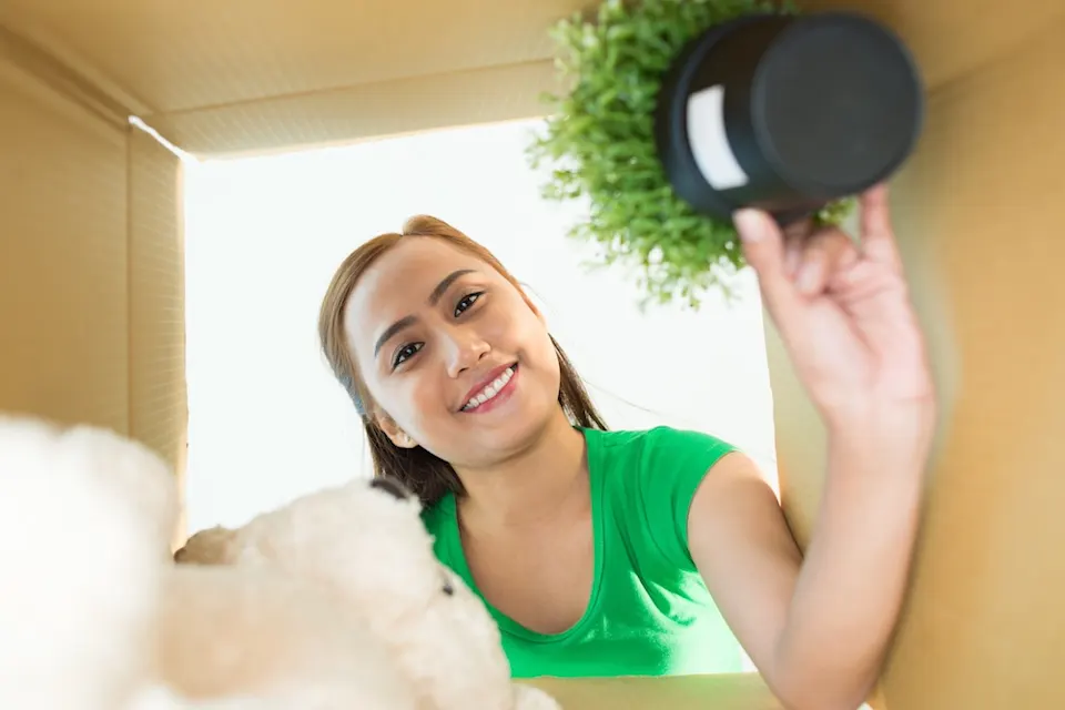 Smiling woman unpacking items from a moving box, including a small plant and a teddy bear.