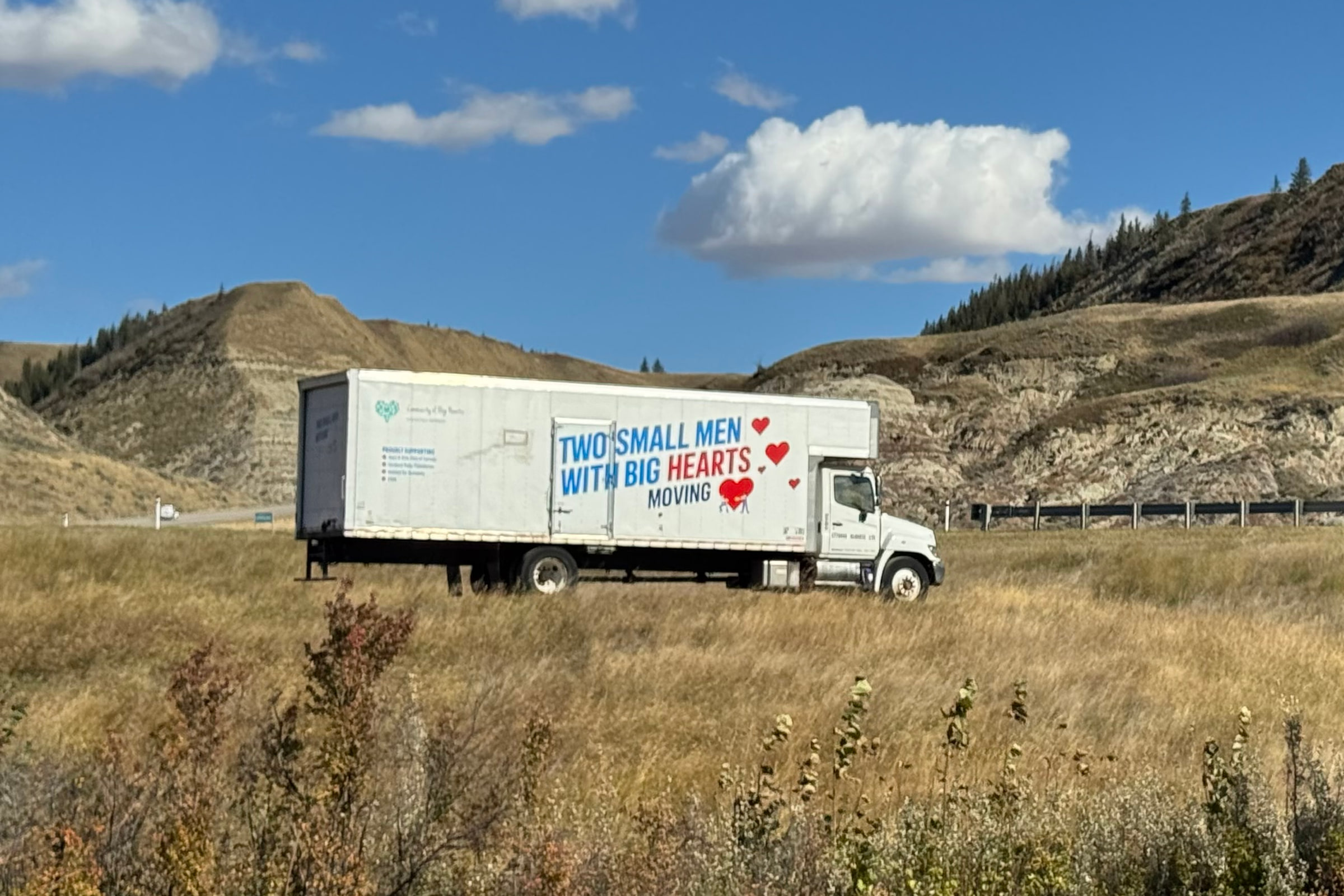 Two Small Men With Big Hearts moving truck parked on a grassy roadside below Alberta badlands hills on a sunny day