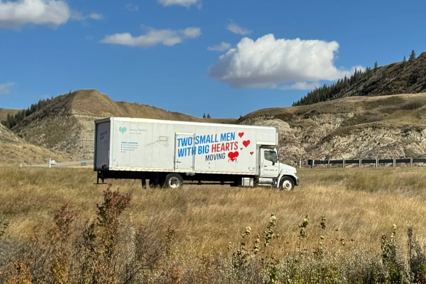 Two Small Men With Big Hearts moving truck parked on a grassy roadside below Alberta badlands hills on a sunny day