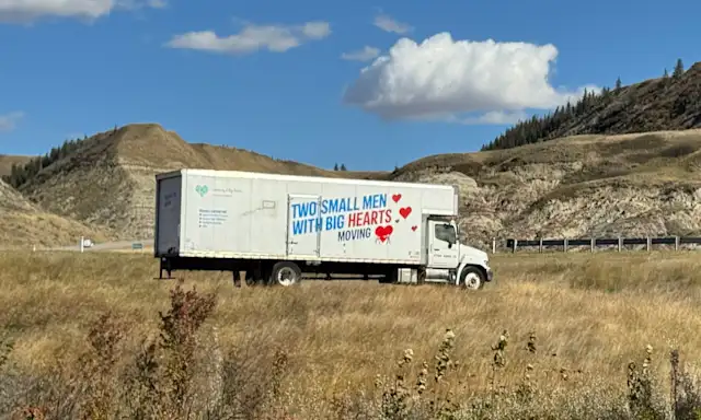 Two Small Men With Big Hearts moving truck parked on a grassy roadside below Alberta badlands hills on a sunny day