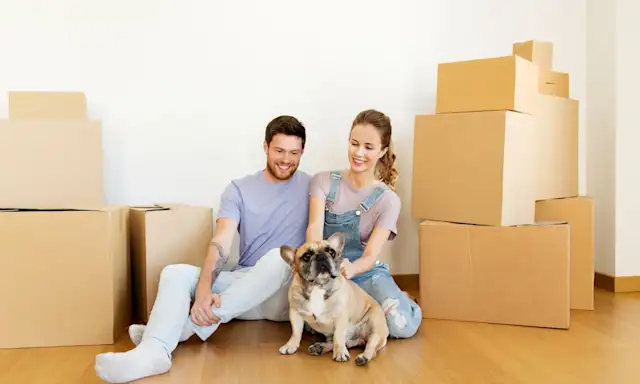 Couple sitting on the floor with their dog, surrounded by moving boxes.