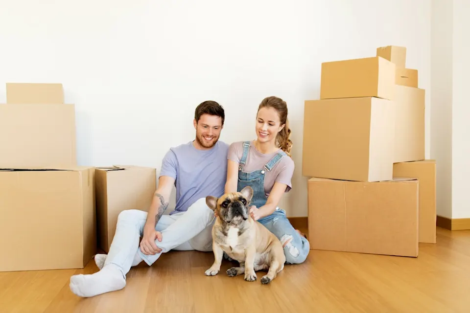 Couple sitting on the floor with their dog, surrounded by moving boxes.
