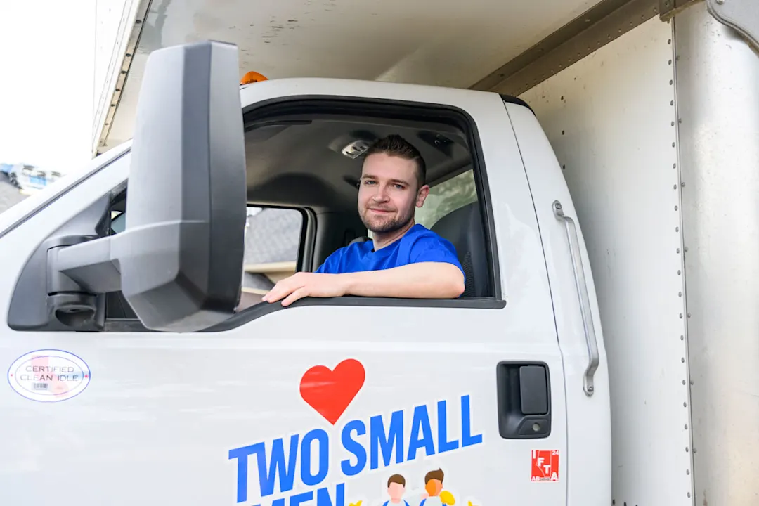 A mover sits in the driver's seat of a "Two Small Men with Big Hearts" moving truck, smiling through the open window. The company logo with a red heart is visible on the door.