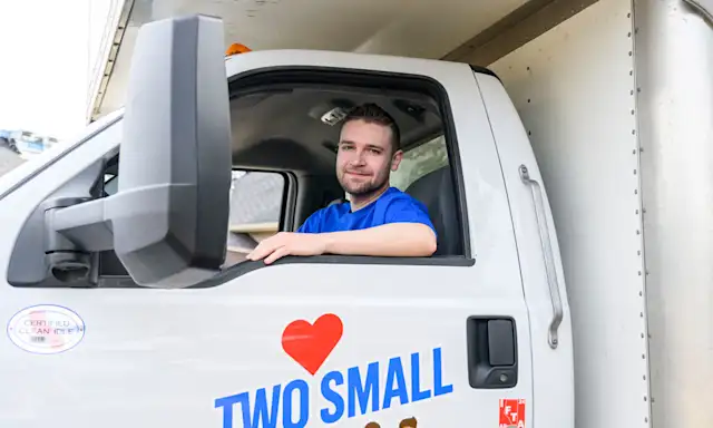 A mover sits in the driver's seat of a "Two Small Men with Big Hearts" moving truck, smiling through the open window. The company logo with a red heart is visible on the door.