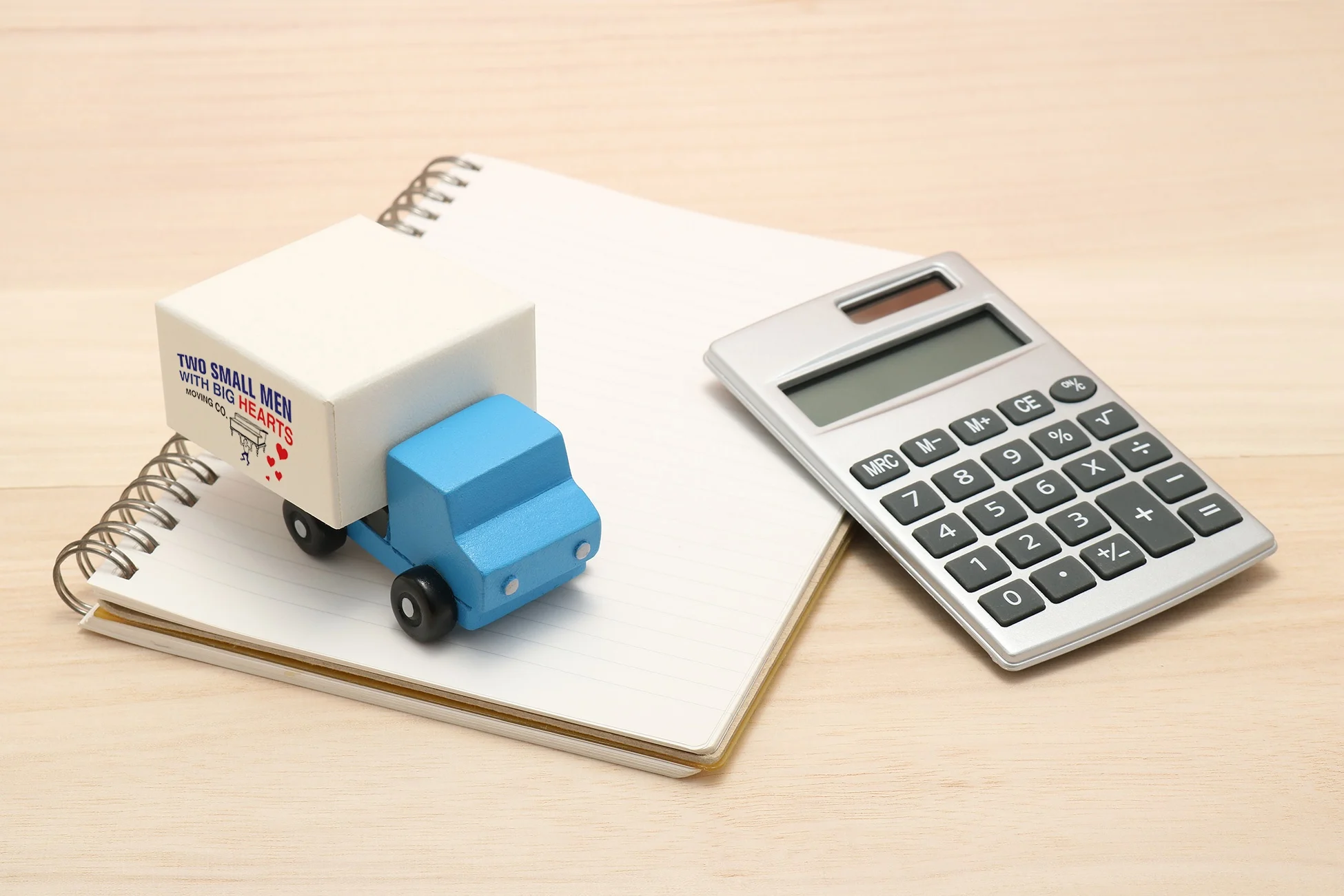 A miniature "Two Small Men with Big Hearts" moving truck sits on a notebook next to a calculator, symbolizing moving cost calculations.