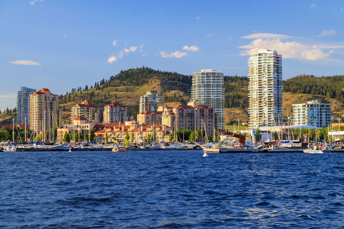 Waterfront view of Kelowna with high-rise buildings, marina, and hills in the background on a sunny day.