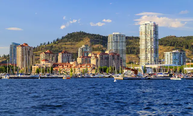 Waterfront view of Kelowna with high-rise buildings, marina, and hills in the background on a sunny day.