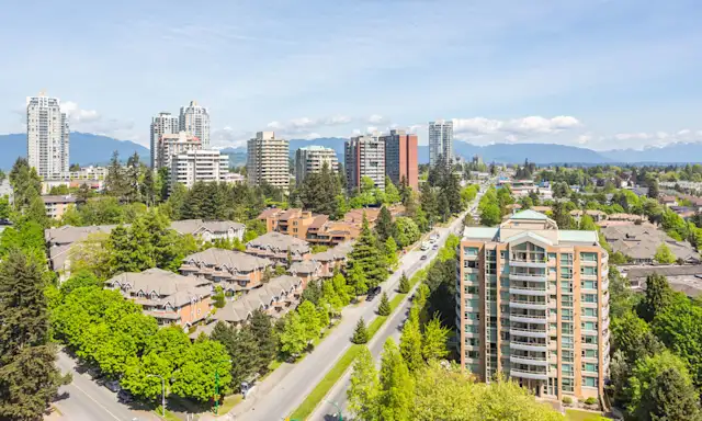 Aerial view of residential and commercial buildings in Burnaby, surrounded by trees with mountains in the background.