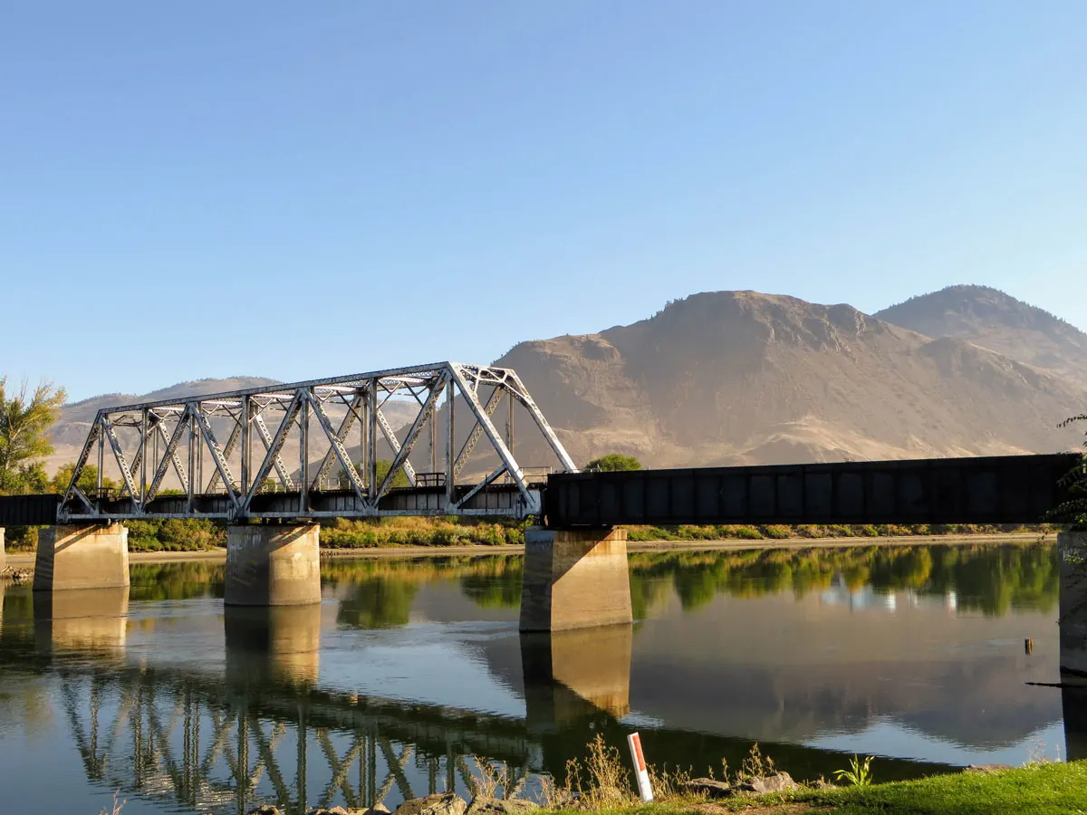 Bridge over a calm river with mountains in the background on a sunny day in Kamloops.