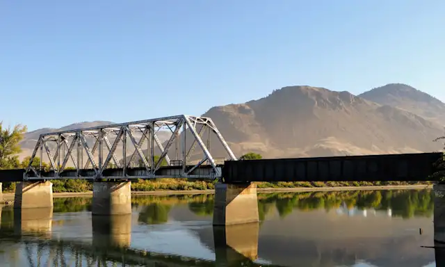 Bridge over a calm river with mountains in the background on a sunny day in Kamloops.