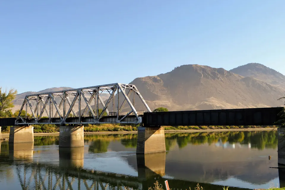 Bridge over a calm river with mountains in the background on a sunny day in Kamloops.
