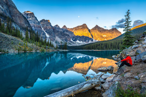 Person sitting by the shore of Moraine Lake with a stunning reflection of the Rocky Mountains in Banff National Park, Alberta, at sunrise.