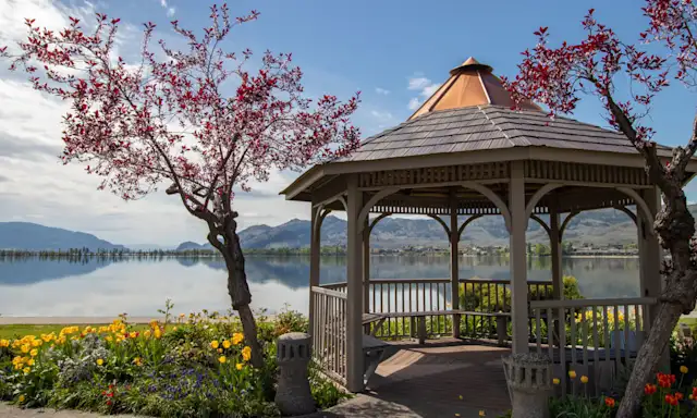 Gazebo by a lake in Osoyoos, with blooming trees and flowers, surrounded by mountains under a clear sky.